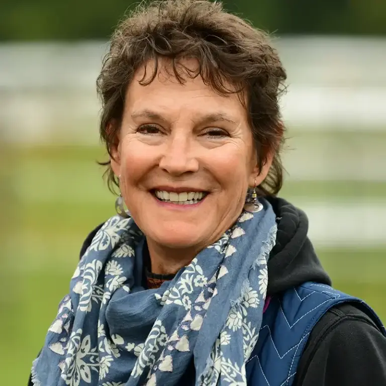 A woman with short, curly brown hair smiles outdoors. She is wearing a blue scarf with a white floral pattern, a dark jacket, and earrings. The background is blurred greenery, suggesting a park or garden setting.