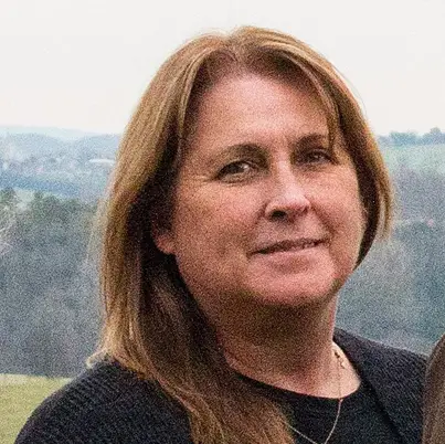 A woman with straight, light brown hair and a black sweater stands outdoors, with a blurred background of trees and grassy fields. She is looking at the camera with a slight smile.