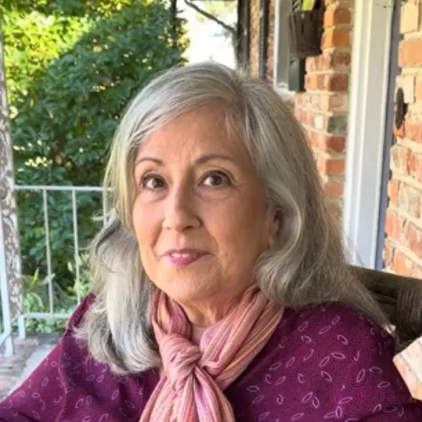 An older woman with long gray hair and a pink scarf sits on a porch. She is wearing a maroon top with small patterns and is looking at the camera, with greenery and a brick wall in the background.