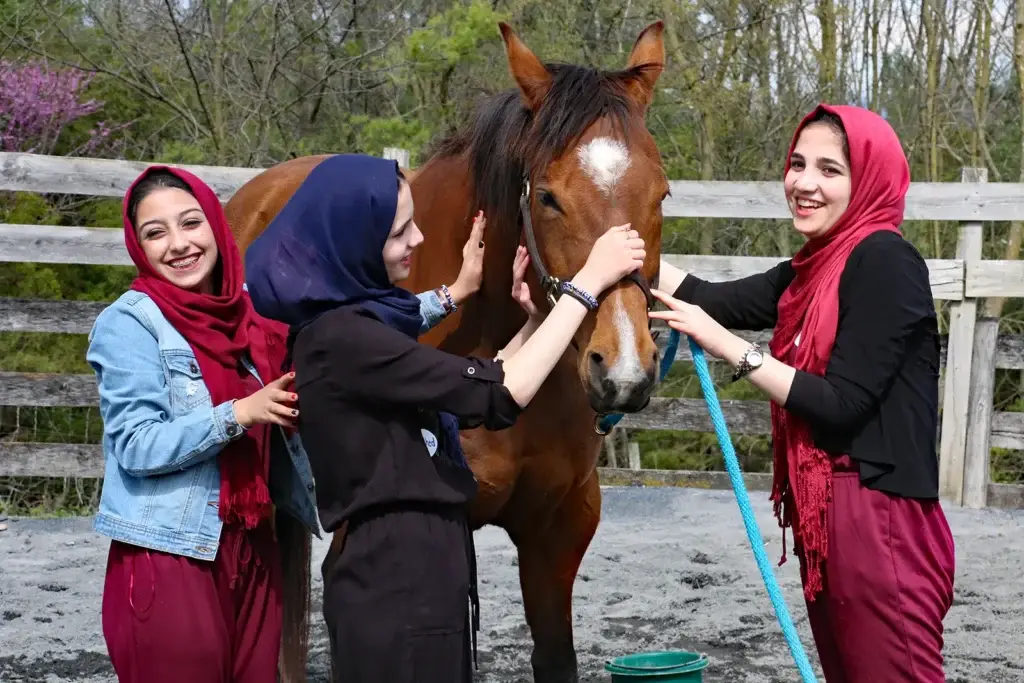 Three smiling young women wearing hijabs and matching maroon outfits stand beside a brown horse, gently petting it, outdoors by a wooden fence with trees in the background.