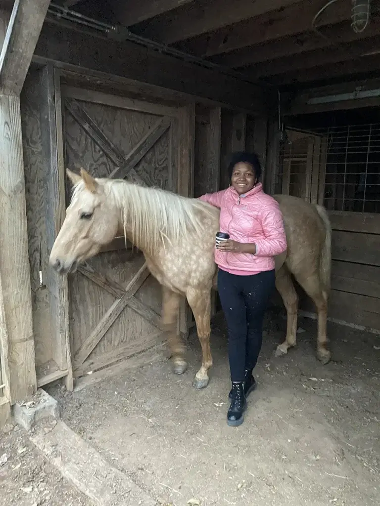 A person wearing a pink jacket and black pants stands in a wooden stable, smiling and holding a cup, with one hand resting on a light-colored horse beside them.