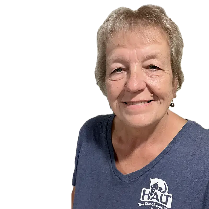 An older woman with short light hair smiles at the camera. She is wearing a blue V-neck shirt with a logo that says HALT and features a dog and the words Healing Animals with Love & Trust.