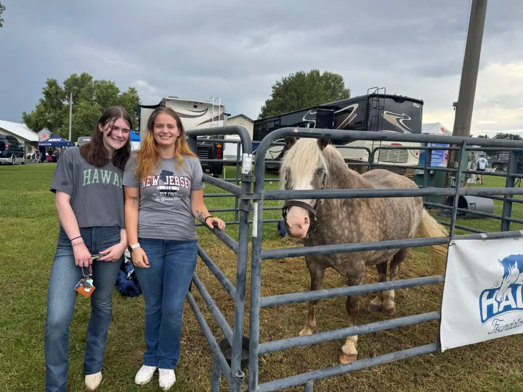 Two young women stand smiling next to a fenced-in horse at an outdoor event. They wear casual clothes and hold drinks. RVs, a tent, and a cloudy sky are visible in the background.