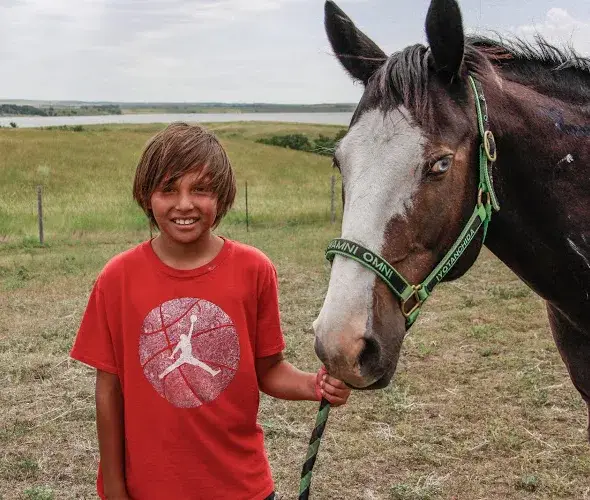 A smiling child in a red shirt with a basketball logo stands outside, holding the reins of a dark brown horse with a white stripe on its face. They are in a grassy field under a cloudy sky.