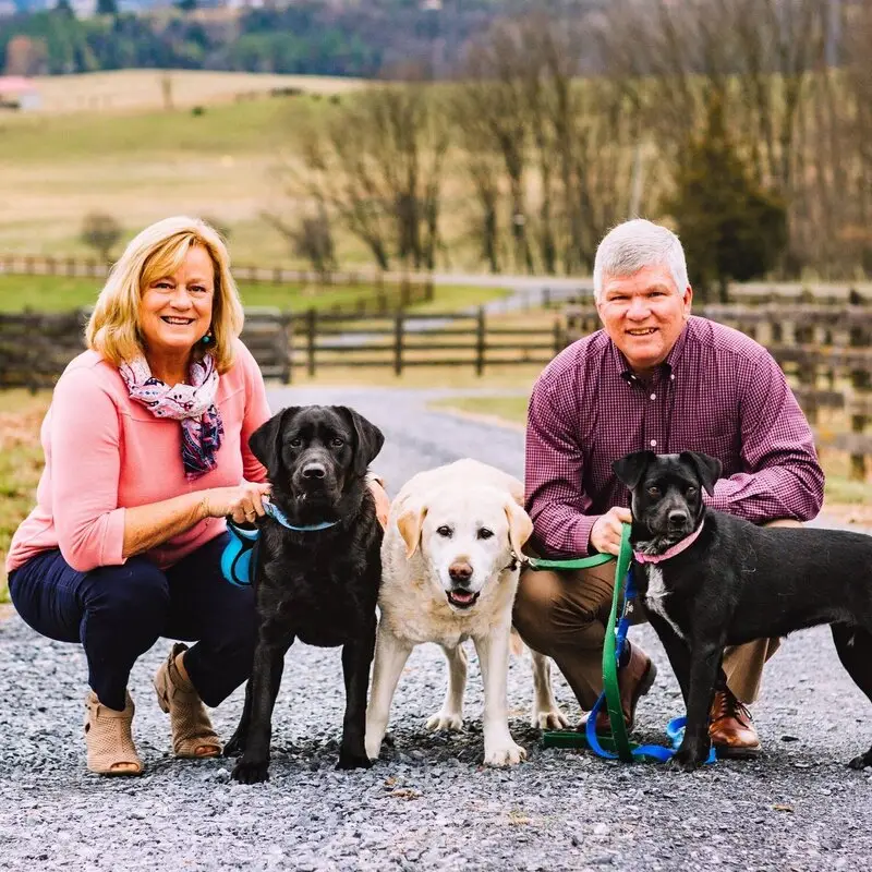 A smiling man and woman kneel on a gravel path with three dogs—two black and one yellow—on a leash. Fields, fences, and bare trees are visible in the background.