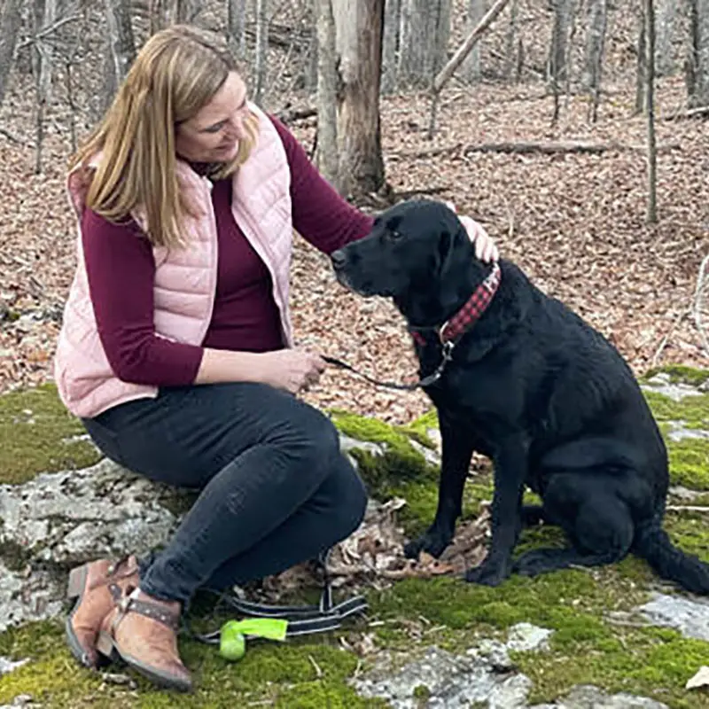 A woman in a pink vest sits on a mossy rock in a forest, smiling and petting a black dog wearing a red collar. A green tennis ball and a dog leash lie on the ground beside them.