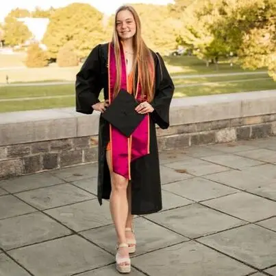 A young woman in a graduation gown and sash stands outdoors on a paved area, holding her graduation cap and smiling. Trees and grass are visible in the background.