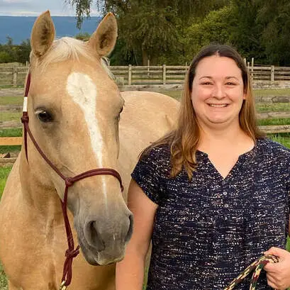 A smiling woman stands outdoors next to a light brown horse with a white blaze on its face, holding the horses lead rope. A wooden fence and trees are visible in the background.