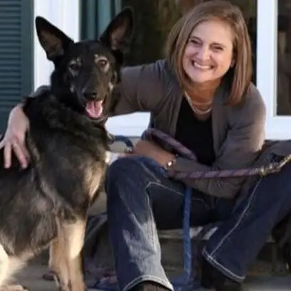 A woman sitting on steps, smiling and hugging a large black-and-tan German Shepherd dog. The dog is standing and facing the camera, both appear happy and relaxed.