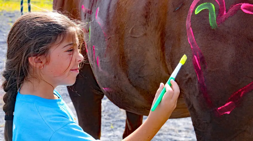 A young girl with braided hair and a blue shirt paints pink and green designs on the side of a brown horse outdoors.
