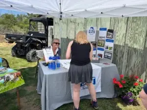 A woman stands at an outdoor booth with brochures and blue bottles, speaking to another woman seated behind the table under a white tent. A wooden fence, utility vehicle, and flowers are visible in the background.