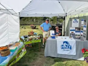Two people at an outdoor booth under a white canopy promote the HALT Foundation. The table displays brochures, water bottles, and a donation box. Another table holds snacks and fruit, with farm fencing visible in the background.