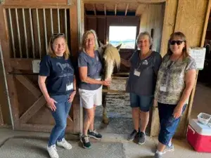 Four smiling women stand in a barn doorway with a white horse, posing for a photo. The women wear casual clothes and name tags. The barn interior is wooden, and sunlight streams in from outside.